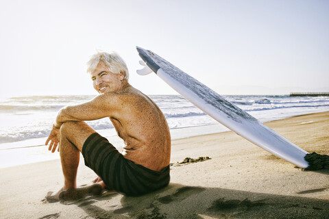 Älterer kaukasischer Mann am Strand sitzend mit Surfbrett, lizenzfreies Stockfoto