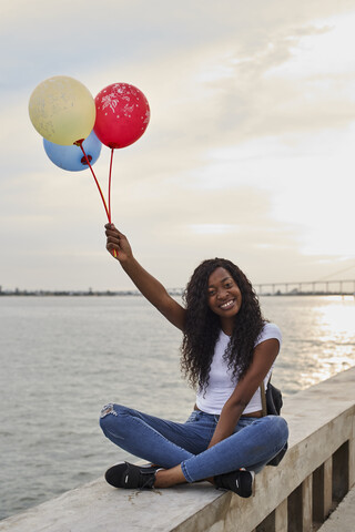 Mocambique, Maputo, Porträt einer glücklichen jungen Frau mit drei Luftballons vor dem Meer sitzend, lizenzfreies Stockfoto