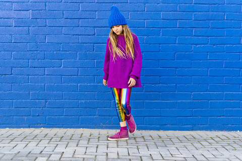 Portrait of girl wearing blue cap and oversized pink pullover standing in front of blue wall looking down stock photo