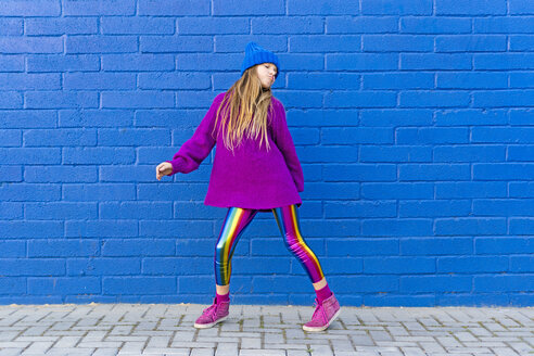 Porträt eines Mädchens mit blauer Mütze und übergroßem rosa Pullover, das vor einer blauen Wand steht und tanzt - ERRF01199