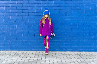 Mädchen mit blauer Mütze und übergroßem rosa Pullover steht vor einer blauen Wand und hört mit Kopfhörern Musik - ERRF01200
