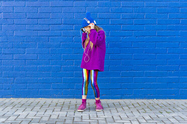 Girl wearing blue cap and oversized pink pullover standing in front of blue wall listening music with headphones - ERRF01201