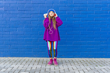Girl wearing blue cap and oversized pink pullover standing in front of blue wall listening music with headphones - ERRF01202