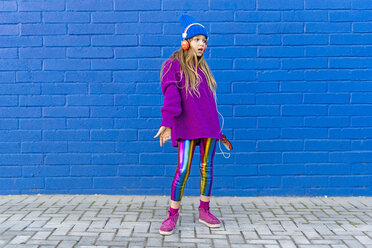 Girl wearing blue cap and oversized pink pullover standing in front of blue wall listening music with headphones - ERRF01203