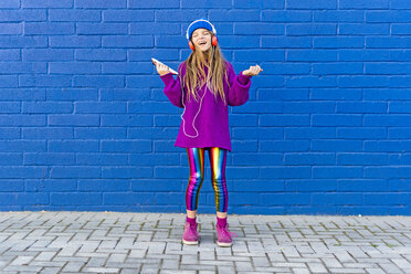 Girl wearing blue cap and oversized pink pullover standing in front of blue wall listening music with headphones - ERRF01204