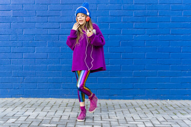 Singing girl dancing in front of blue wall while listening music with headphones - ERRF01205