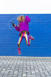Mädchen mit Kopfhörern und Smartphone, das vor einer blauen Wand in die Luft springt - ERRF01207