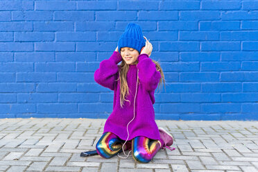 Mädchen mit blauer Mütze und übergroßem rosa Pullover hört Musik mit Kopfhörern vor einer blauen Wand - ERRF01209