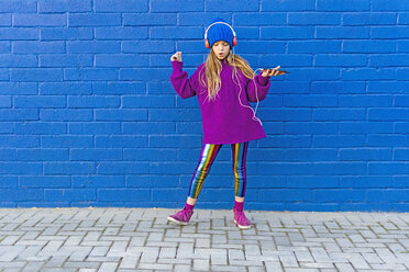 Girl wearing blue cap and oversized pink pullover listening music with headphones in front of blue wall - ERRF01210