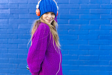 Portrait of smiling girl with headphones wearing blue cap and pink pullover in front of blue wall - ERRF01212