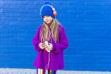 Girl wearing blue cap and oversized pink pullover listening music with headphones in front of blue wall - ERRF01213