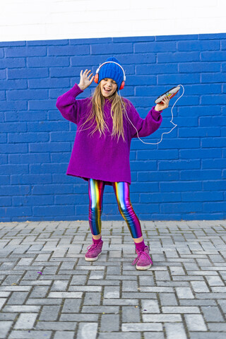 Singendes Mädchen, das vor einer blauen Wand tanzt, während es mit Kopfhörern Musik hört, lizenzfreies Stockfoto