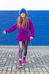 Happy girl dancing in front of blue wall while listening music with headphones and smartphone - ERRF01218