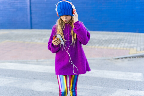 Mädchen mit blauer Mütze und übergroßem rosa Pullover hört Musik mit Kopfhörern und Smartphone, lizenzfreies Stockfoto