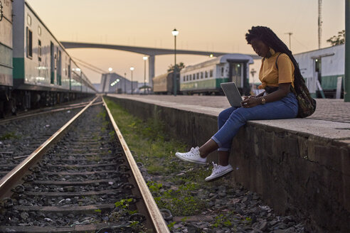 Young woman sitting on platform at the train station using laptop - VEGF00117