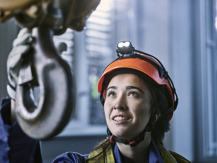 Junge Industriearbeiterin mit Schutzhelm nimmt den Haken eines Hallenkrans - CVF01125