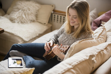 Smiling woman sitting on sofa playing ukulele - BLEF01820