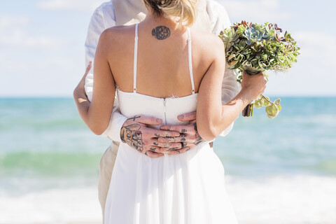 Caucasian bride and groom with tattoos hugging on beach stock photo