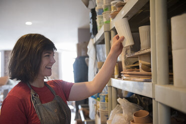 Smiling Caucasian woman placing cup on shelf in workshop - BLEF01884