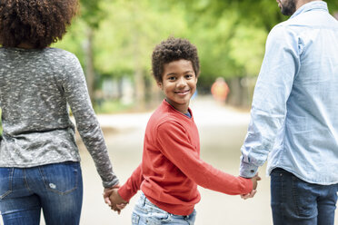 Happy boy holding parent's hands, walking in a park - JSMF01057