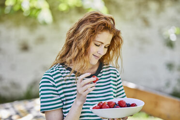 Porträt einer lächelnden rothaarigen jungen Frau mit einer Schale voller Beeren im Garten - FMKF05662