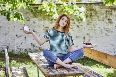 Portrait of young woman sitting on wooden table in garden holding bowl of berries and glass of yoghurt - FMKF05663