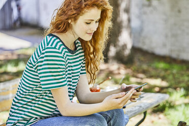 Smiling redheaded young woman sitting on bench in the garden looking at cell phone - FMKF05669