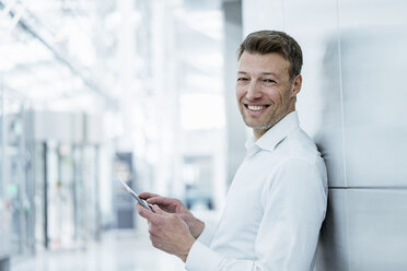 Porträt eines lächelnden Geschäftsmannes, der an einer Wand lehnt und ein Mobiltelefon hält - DIGF06837