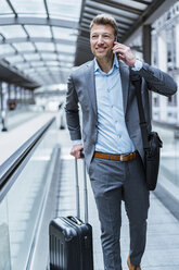 Smiling businessman with baggage and cell phone on moving walkway - DIGF06904