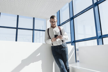 Businessman using cell phone at the window in modern office - AHSF00213