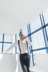 Businessman talking on cell phone at the window in modern office - AHSF00214