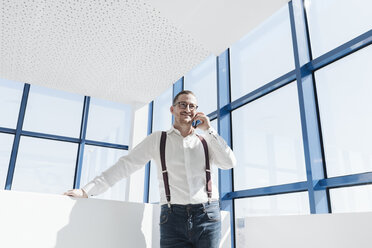 Businessman talking on cell phone at the window in modern office - AHSF00215