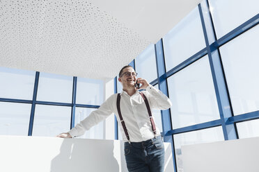 Glücklicher Geschäftsmann, der am Fenster in einem modernen Büro mit seinem Handy telefoniert - AHSF00216