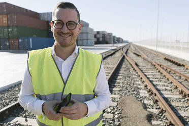 Portrait of smiling man on railway tracks in front of cargo containers with cell phone - AHSF00223