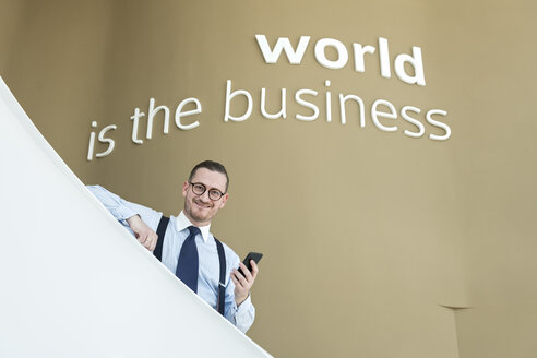 Porträt eines selbstbewussten Geschäftsmannes mit Mobiltelefon auf einer Treppe in einem modernen Büro - AHSF00249