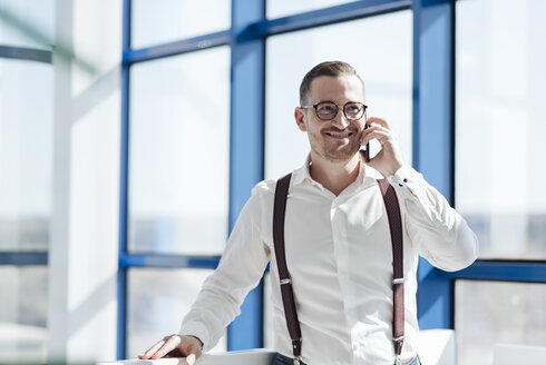 Lächelnder Geschäftsmann, der am Fenster in einem modernen Büro mit seinem Handy telefoniert - AHSF00260