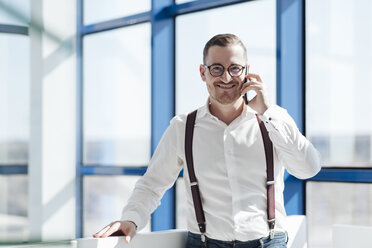 Lächelnder Geschäftsmann, der am Fenster in einem modernen Büro mit seinem Handy telefoniert - AHSF00261