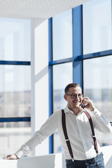 Glücklicher Geschäftsmann, der am Fenster in einem modernen Büro mit seinem Handy telefoniert - AHSF00263