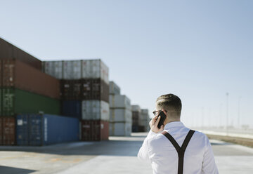 Rückansicht eines Managers, der vor einem Frachtcontainer auf einem Industriegelände mit einem Handy telefoniert - AHSF00272