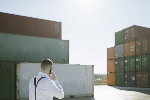 Rückansicht eines Managers, der vor einem Frachtcontainer auf einem Industriegelände mit einem Handy telefoniert - AHSF00273