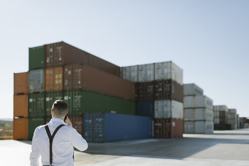 Rückansicht eines Managers, der vor einem Frachtcontainer auf einem Industriegelände mit einem Handy telefoniert - AHSF00274