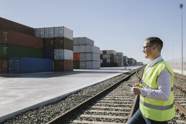 Mann auf Bahngleisen vor Frachtcontainern mit Mobiltelefon in der Hand - AHSF00284