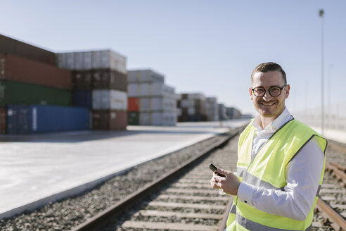 Porträt eines lächelnden Mannes auf Bahngleisen vor Frachtcontainern mit Mobiltelefon - AHSF00289