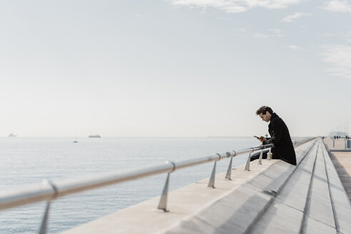 Businessman sitting on quay wall checking cell phone - AFVF02903