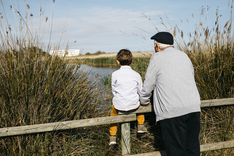 Back view of little boy and his grandfather looking at a lake stock photo