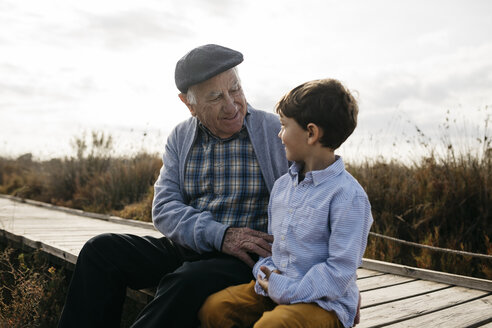 Happy grandfather sitting with his grandson on boardwalk looking at each other - JRFF03186