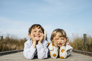 Porträt eines Jungen und seiner kleinen Schwester, die nebeneinander auf der Strandpromenade liegen und lustige Gesichter ziehen - JRFF03188