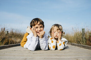Porträt eines Jungen und seiner kleinen Schwester, die nebeneinander auf der Strandpromenade liegen und lustige Gesichter ziehen - JRFF03189