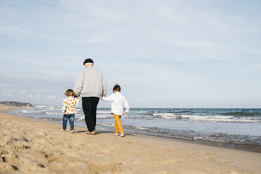 Rückenansicht eines Großvaters, der mit seinen Enkeln Hand in Hand am Strand spazieren geht - JRFF03208