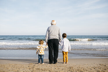 Back view of grandfather standing hand in hand on the beach with his grandchildren watching the sea - JRFF03230